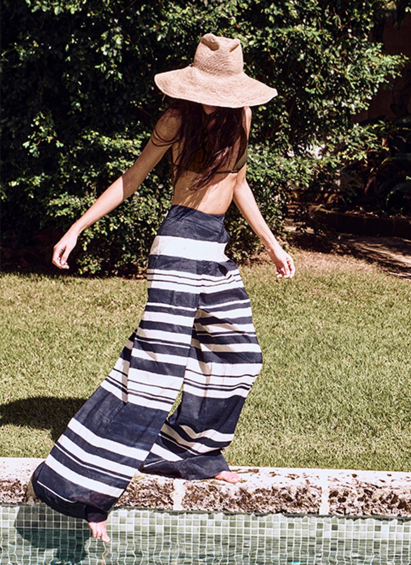 A person wearing a wide-brimmed hat and a striped dress is walking on a grassy area surrounded by lush greenery.