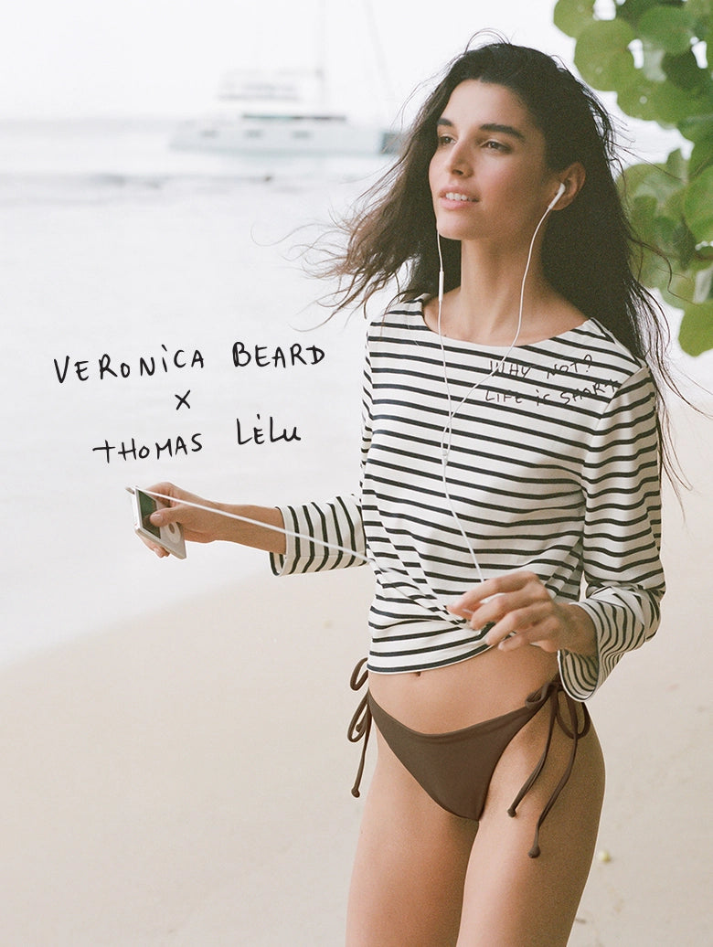 A young woman in a striped shirt and swimsuit stands on a beach, holding a whiteboard with the names "Veronica Beard" and "Thomas Lelu" written on it.