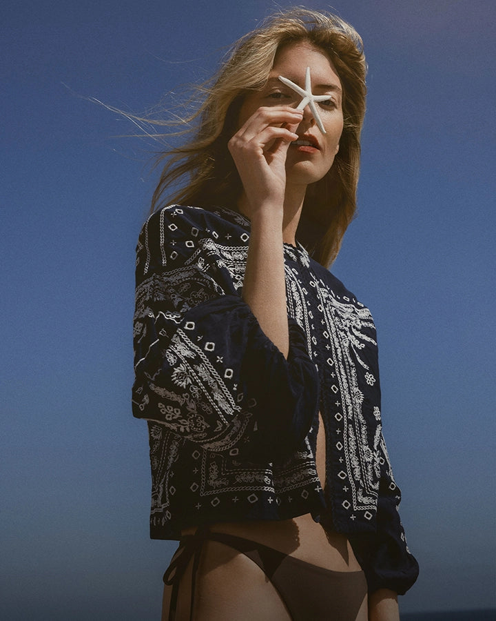A woman in a patterned jacket holds a starfish to her eye against a clear blue sky.