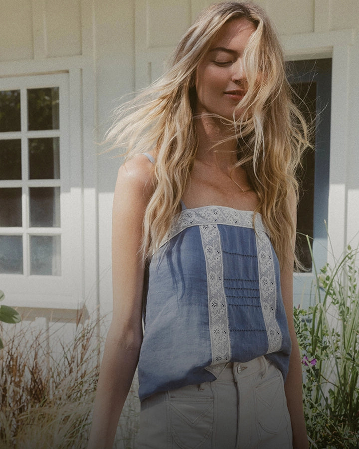 A blonde woman in a blue and white tank top and light-colored pants stands in front of a white building with windows and greenery.