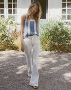 A blonde woman in a blue and white tank top and white flared jeans walks on a gravel path in front of a white building with greenery.