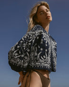 A woman in a patterned jacket and bikini bottoms poses against a clear blue sky.