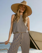 A woman in a striped outfit and wide-brimmed hat holds a surfboard against a backdrop of the ocean and clear sky.