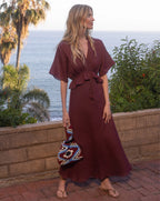 A woman in a maroon dress stands on a brick patio, holding a colorful bag, with palm trees and the ocean in the background.