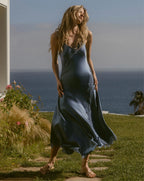 A woman in a blue slip dress stands on a stone path in a grassy yard with the ocean and sky in the background.