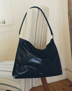 A black leather hobo bag hangs from the back of a striped chair in front of a window and wooden wall.