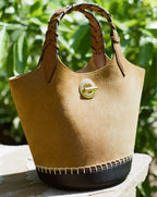 A tan suede bucket bag with a braided handle and dark leather base sits on a stone ledge against a blurred green leafy background.
