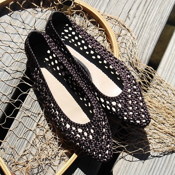 A pair of dark brown, woven crocheted flats rest on a fishing net, which is laid across a weathered wooden dock.