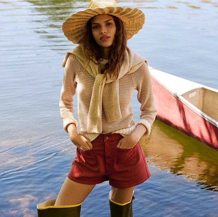 A woman in a straw hat and red shorts stands in shallow water with a canoe in the background.