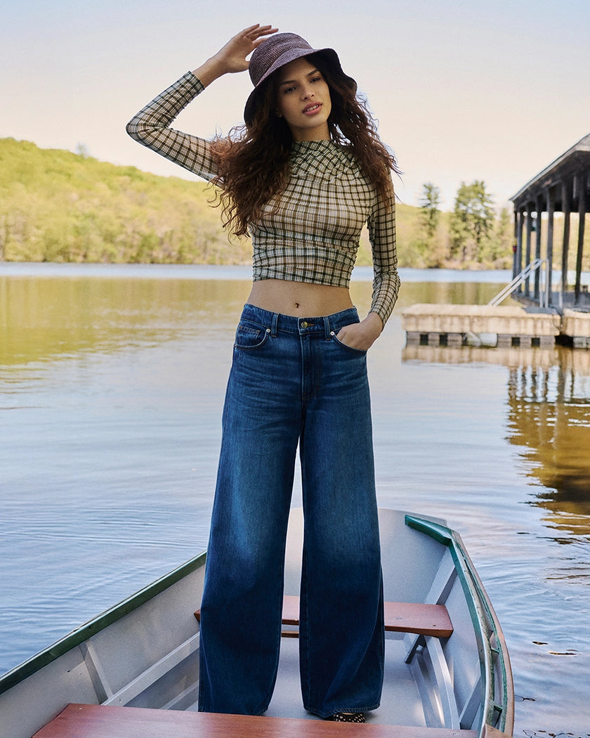 A woman in a checkered top and wide-leg jeans stands in a boat on a lake, with a wooden dock and trees in the background.
