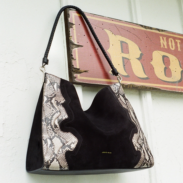 A black suede hobo bag with snakeskin accents hangs in front of a rustic "NOT ROUGH" sign.