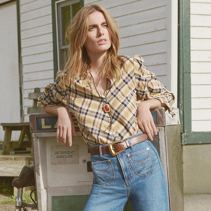 A woman in a plaid shirt and jeans leans against an old gas pump in front of a weathered building.