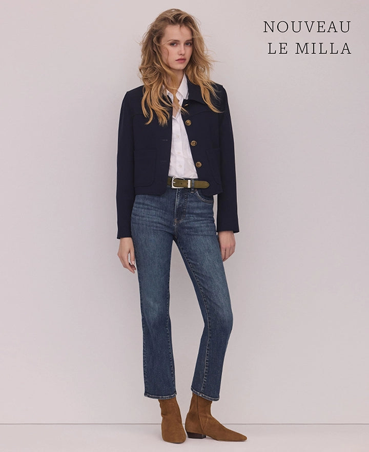 A blonde woman in a navy jacket, white shirt, and blue jeans stands against a plain white background with text in the upper right corner.