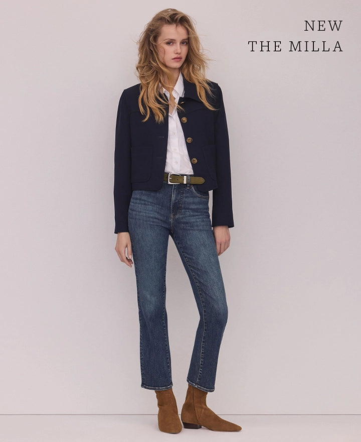 A blonde woman in a navy jacket, white shirt, and blue jeans stands against a plain white background with "NEW THE MILLA" text in the upper right corner.