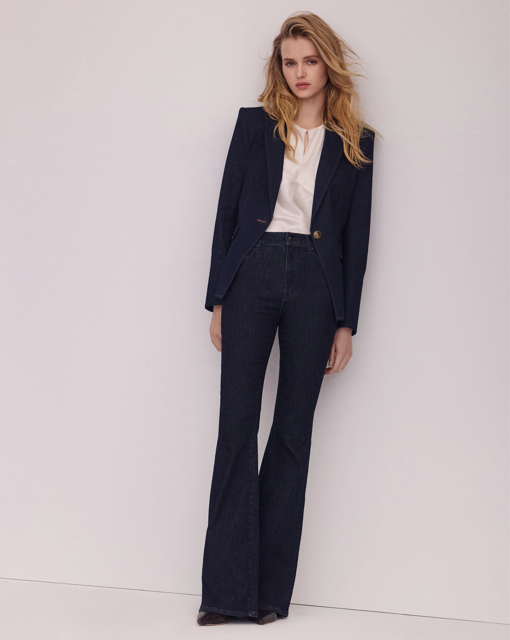 A woman models a dark navy blue tailored suit consisting of a single-breasted blazer and matching high-waisted flare jeans, styled with a cream-colored top against a plain light gray studio background.