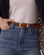 A close-up of a person wearing a brown leather belt with a gold buckle, styled with a white top, blue jeans, a dark blazer, and a leopard-print garment.