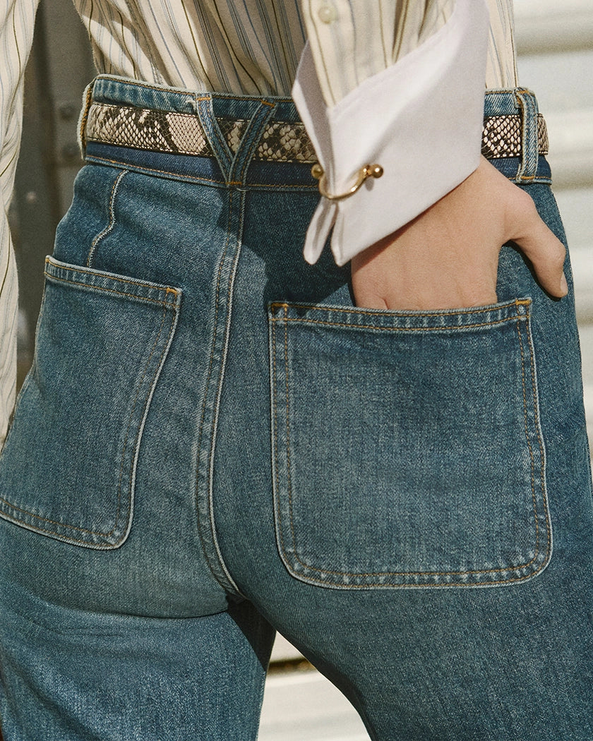 A hand rests in the back pocket of high-waisted denim jeans with a snakeskin belt, against a blurred industrial background.