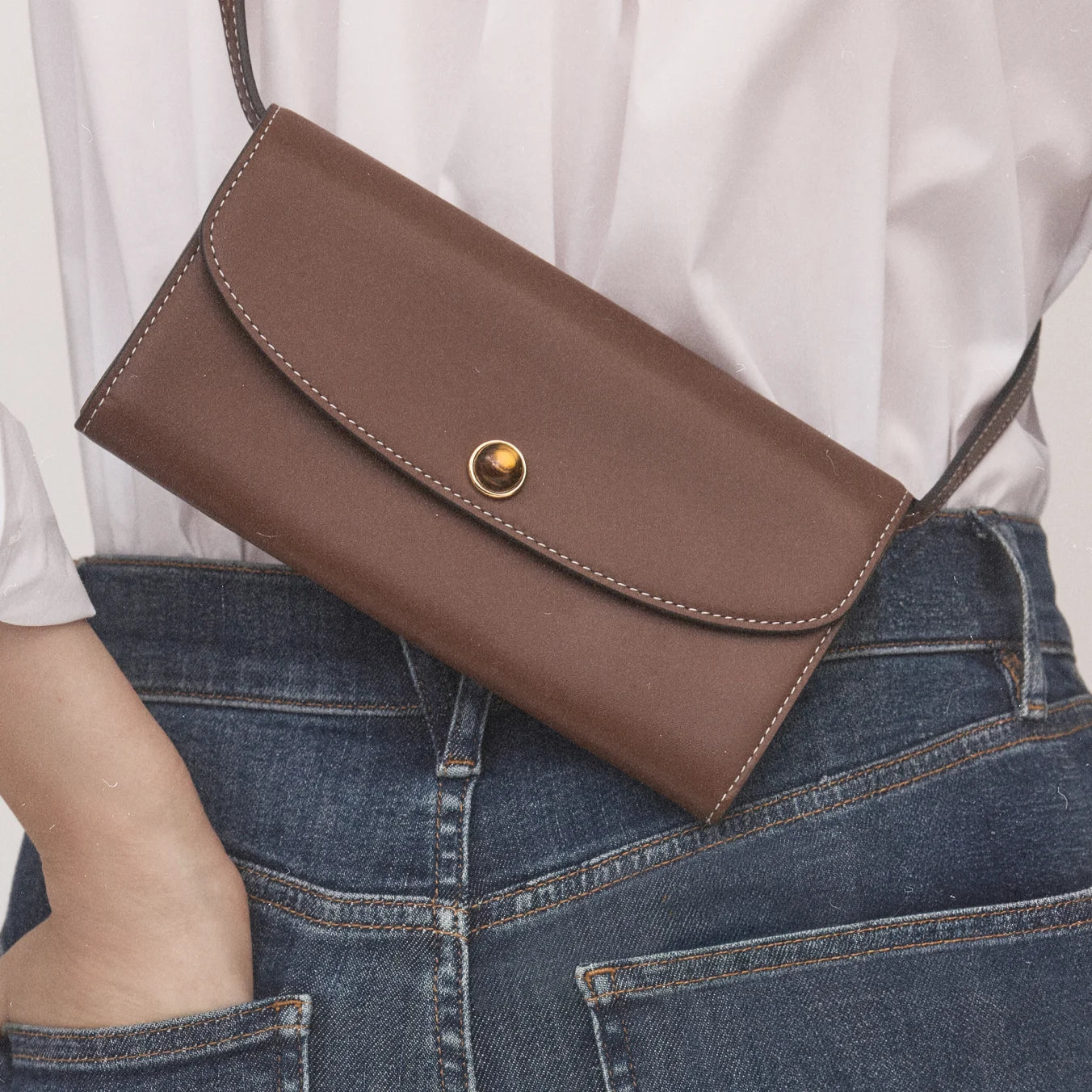 A brown leather clutch with a gold button rests against a person wearing a white shirt and blue jeans.