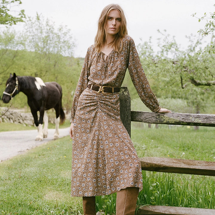 A woman in a patterned dress and boots leans against a wooden fence in the foreground, while a horse stands in a field behind her.