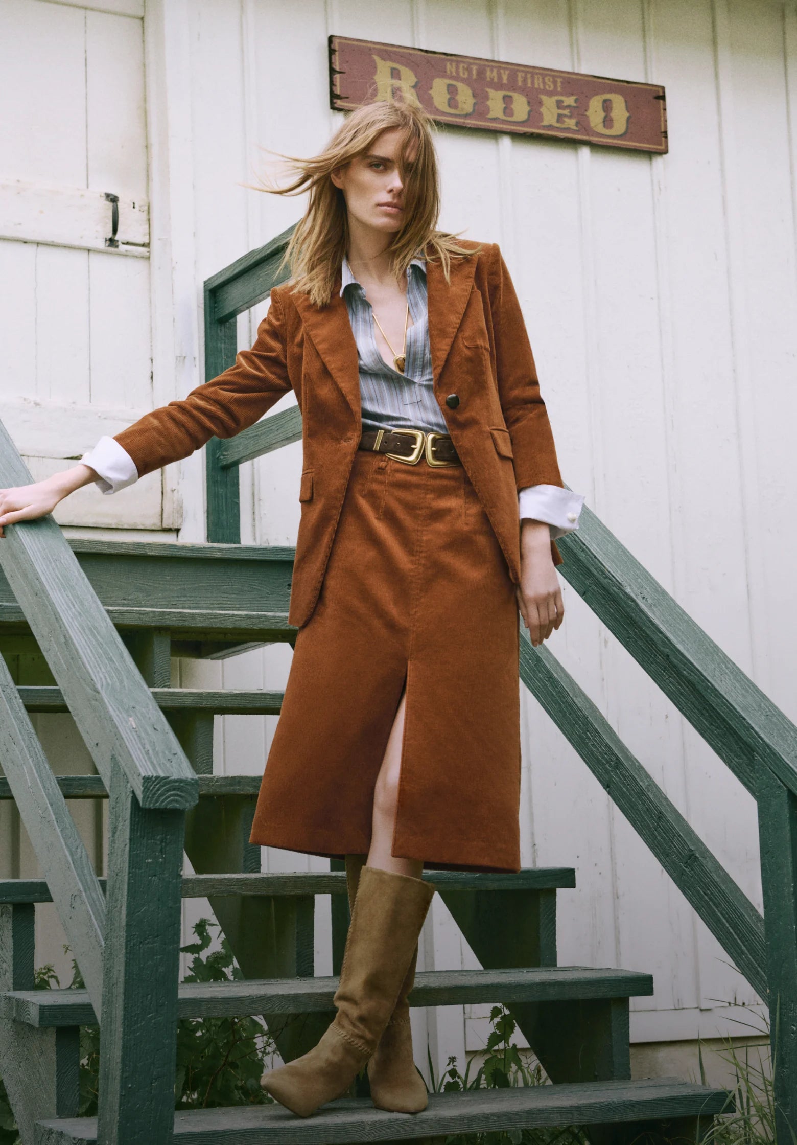 A woman in a brown corduroy suit stands on wooden stairs in front of a white building with a "RODEO" sign.