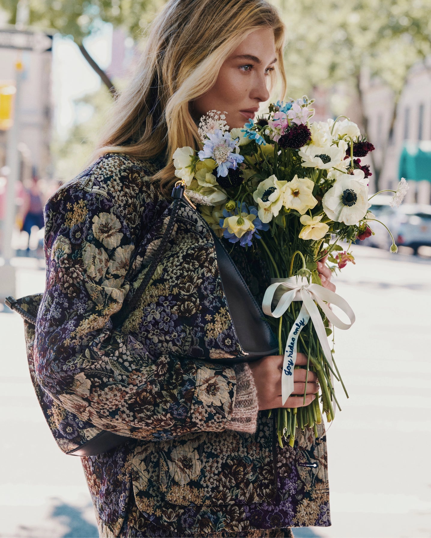 A blonde woman in a floral jacket holds a bouquet of flowers on a sunlit city street.