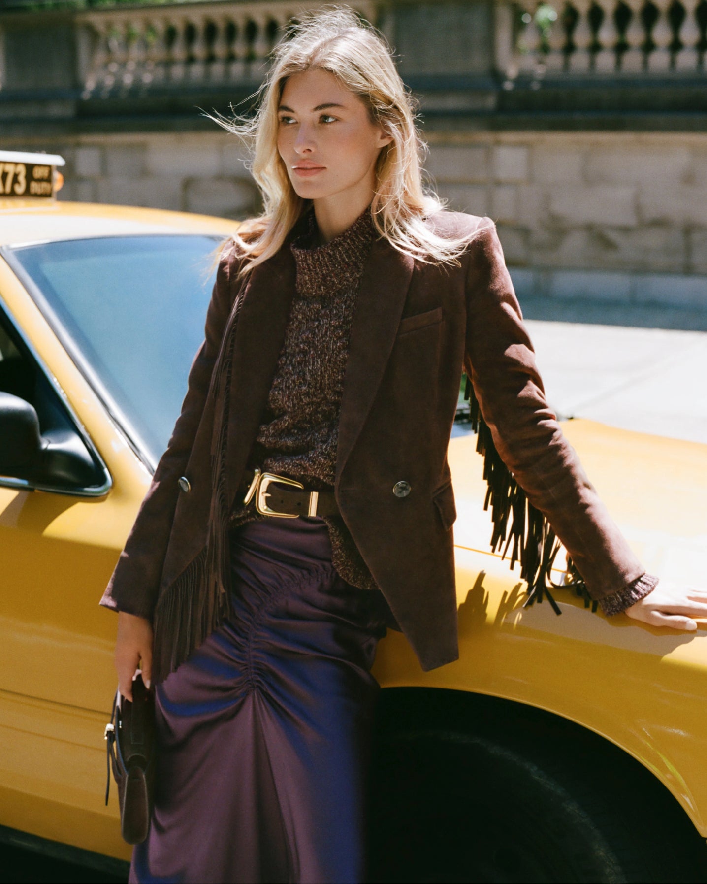 A woman in a brown fringed blazer and purple skirt leans against a yellow taxi, with a blurred city street in the background.