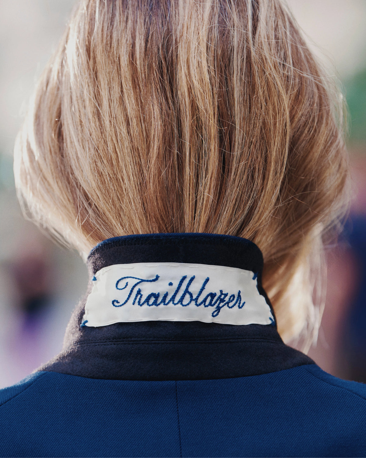 A close-up of a person's blonde hair and the back of a navy jacket with a white collar embroidered with "Trailblazer" is visible against a blurred outdoor background.
