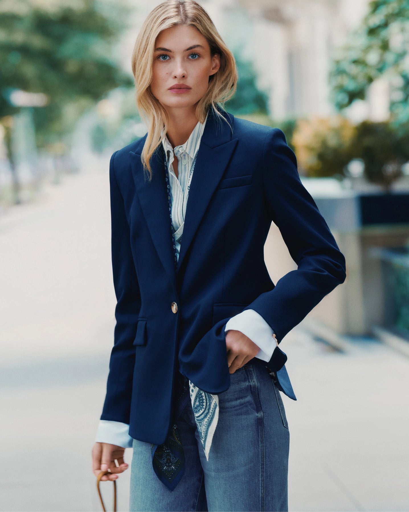 A blonde woman in a navy blazer and jeans stands on a blurred city street.