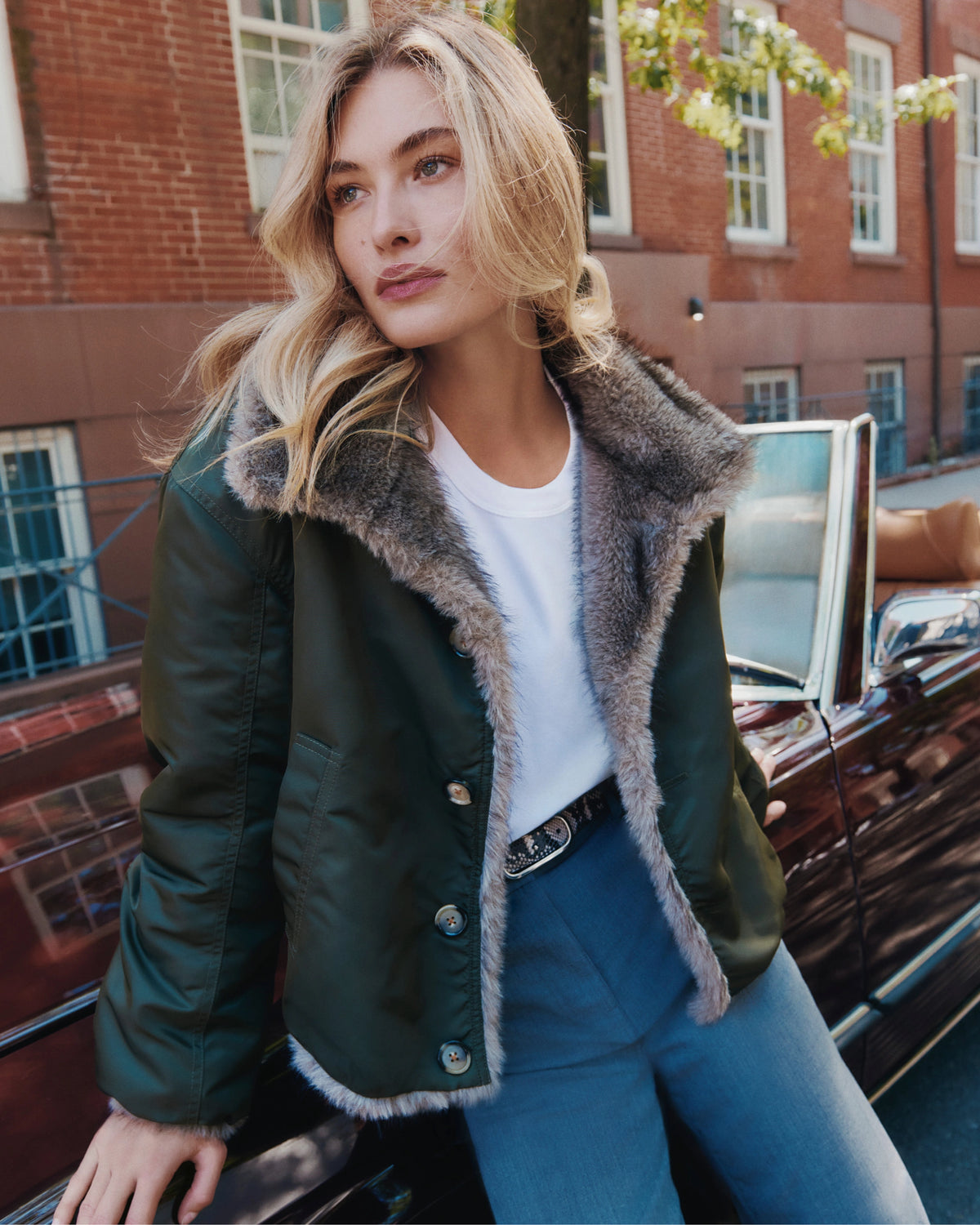 A blonde woman in a green fur-lined jacket leans against a vintage car with a brick building in the background.