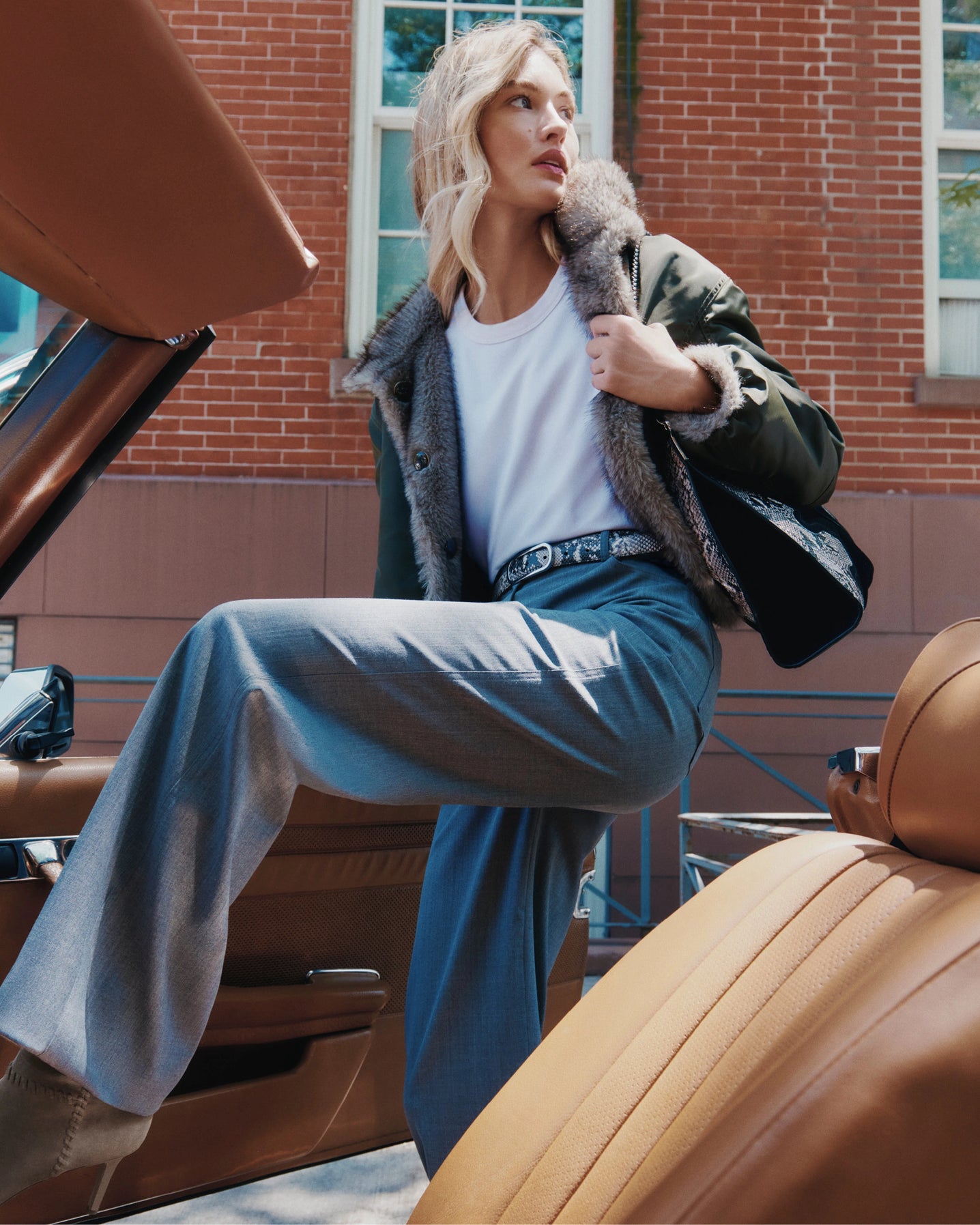 A woman in a stylish outfit steps out of a vintage car with a brick building in the background.