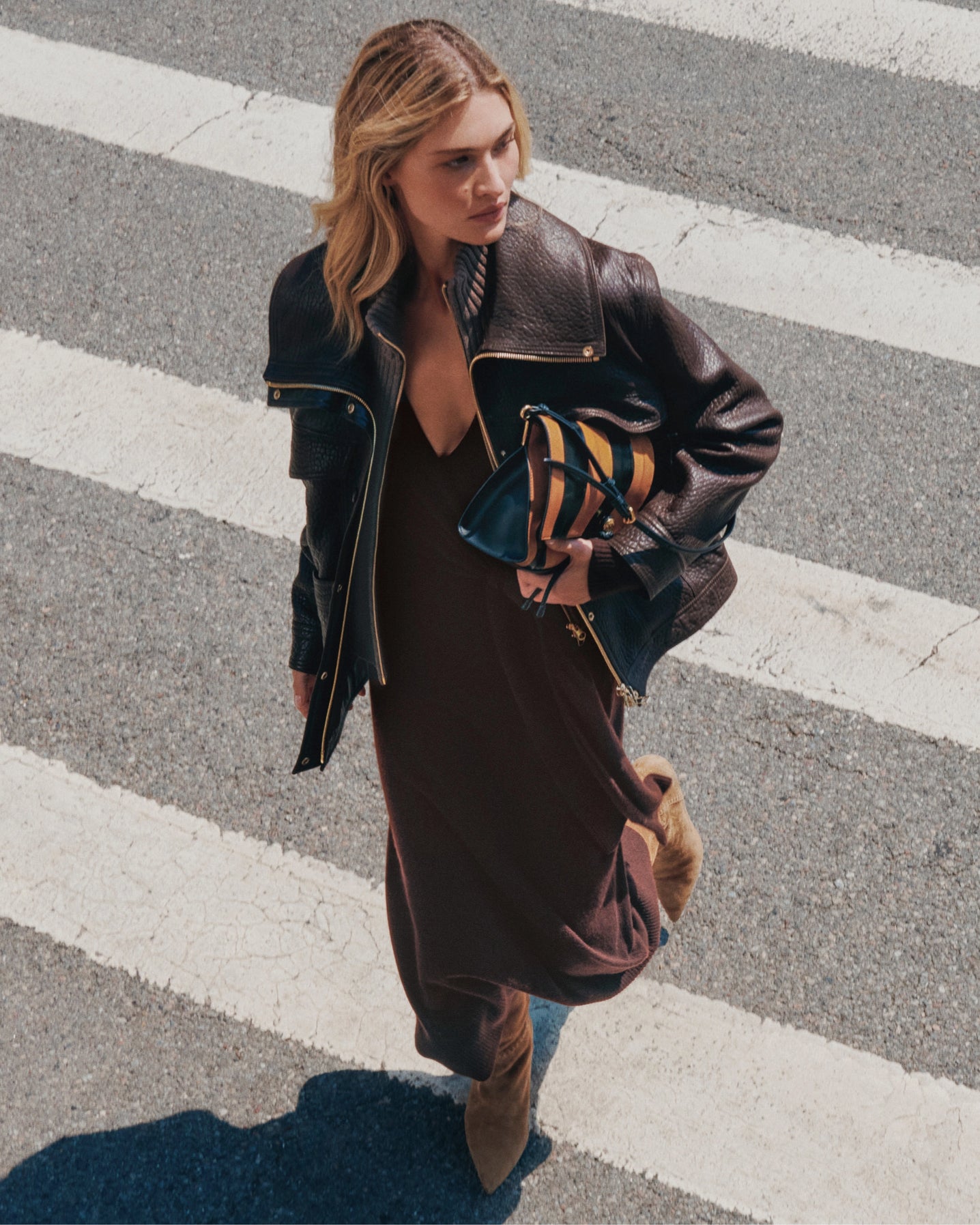 A woman in a brown dress and leather jacket walks across a crosswalk, holding a striped clutch.