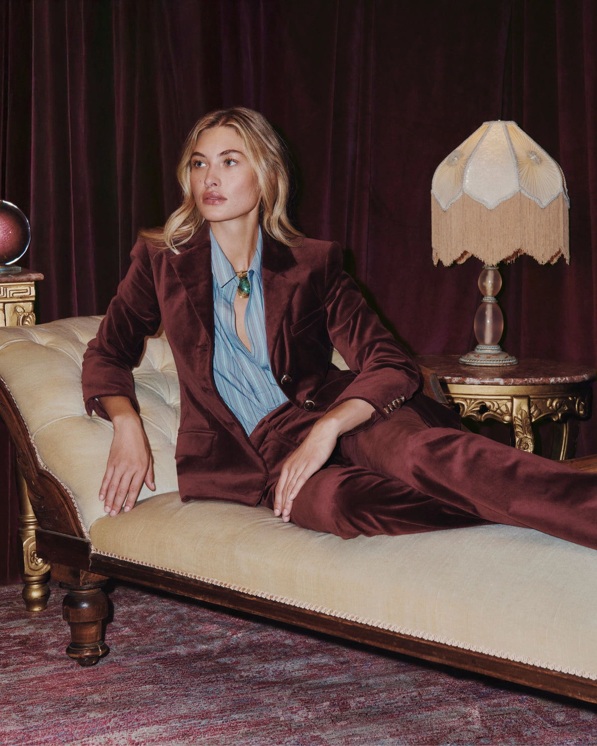 A woman in a burgundy velvet suit reclines on a chaise lounge in front of a dark red curtain and an ornate lamp.