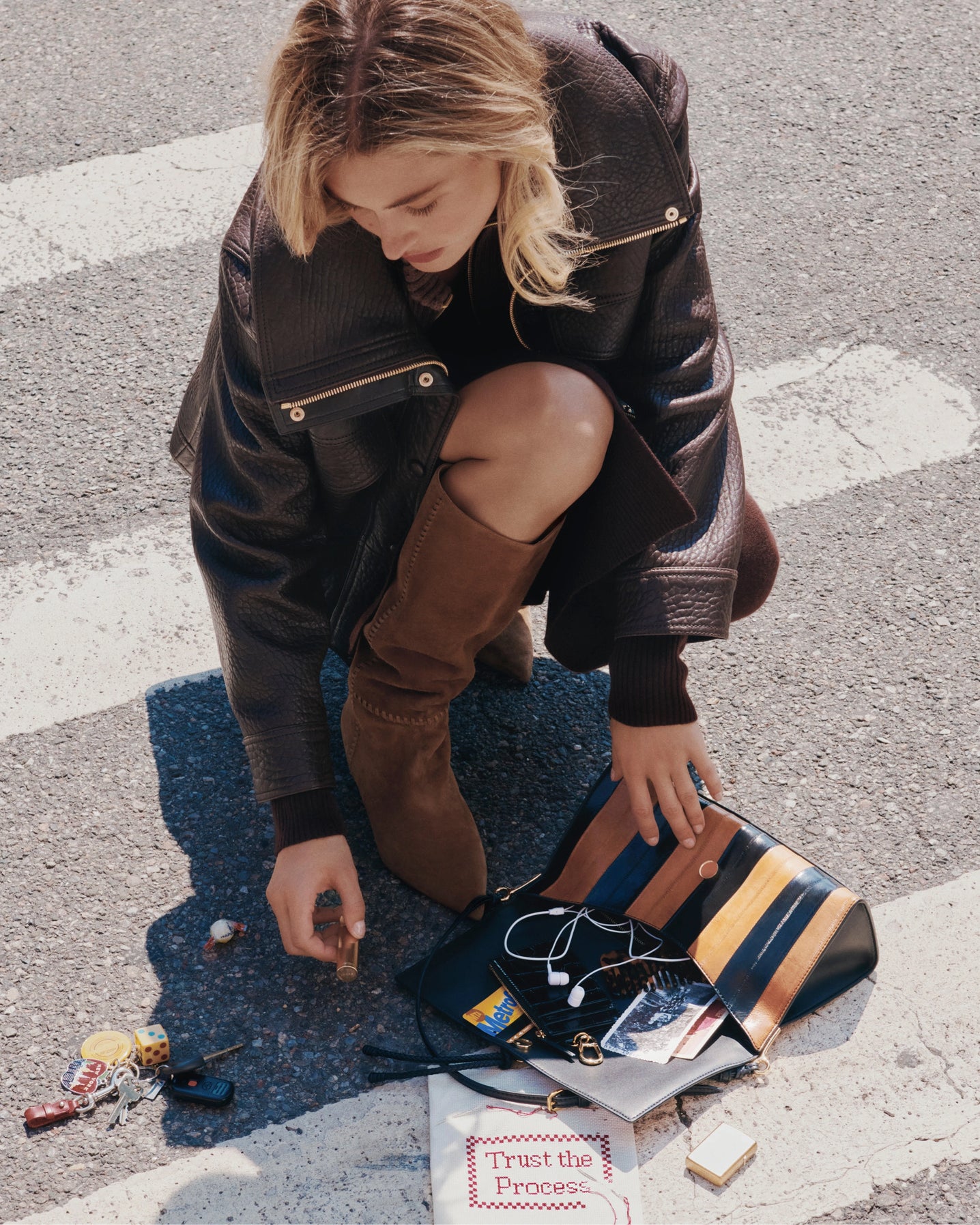A young woman in a leather jacket and brown boots kneels on a crosswalk, reaching into her open purse filled with items, while a shadow stretches across the white stripes of the street.