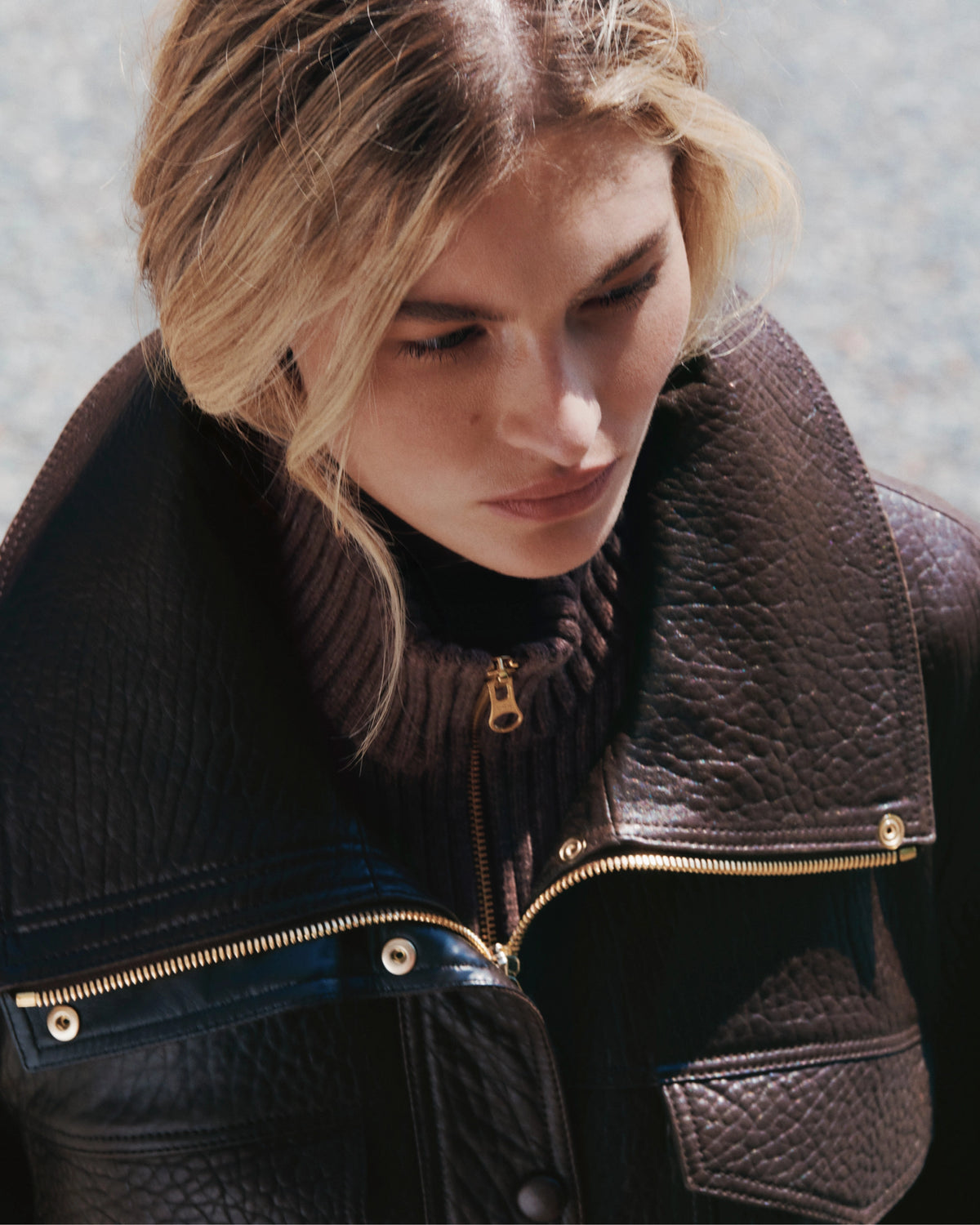 A woman with blonde hair looks down, wearing a dark brown leather jacket over a ribbed turtleneck, with a blurred outdoor background.