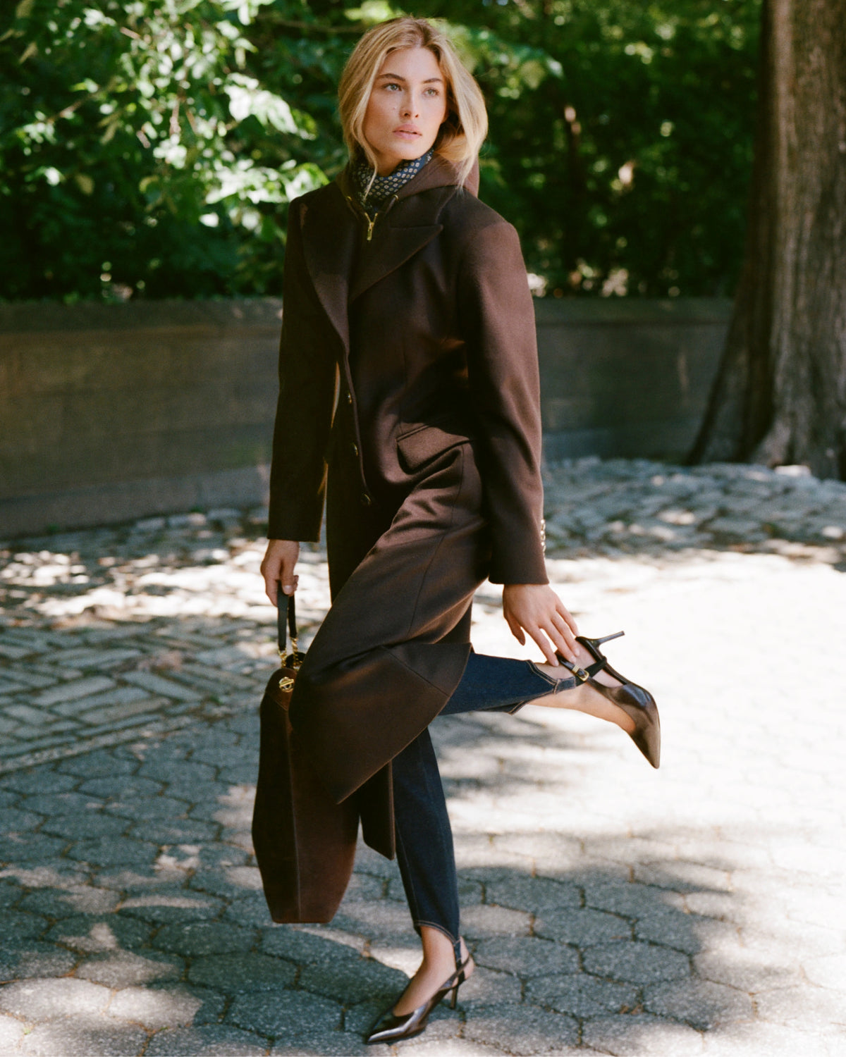 A woman in a brown coat and heels poses on a paved path with lush green trees in the background.