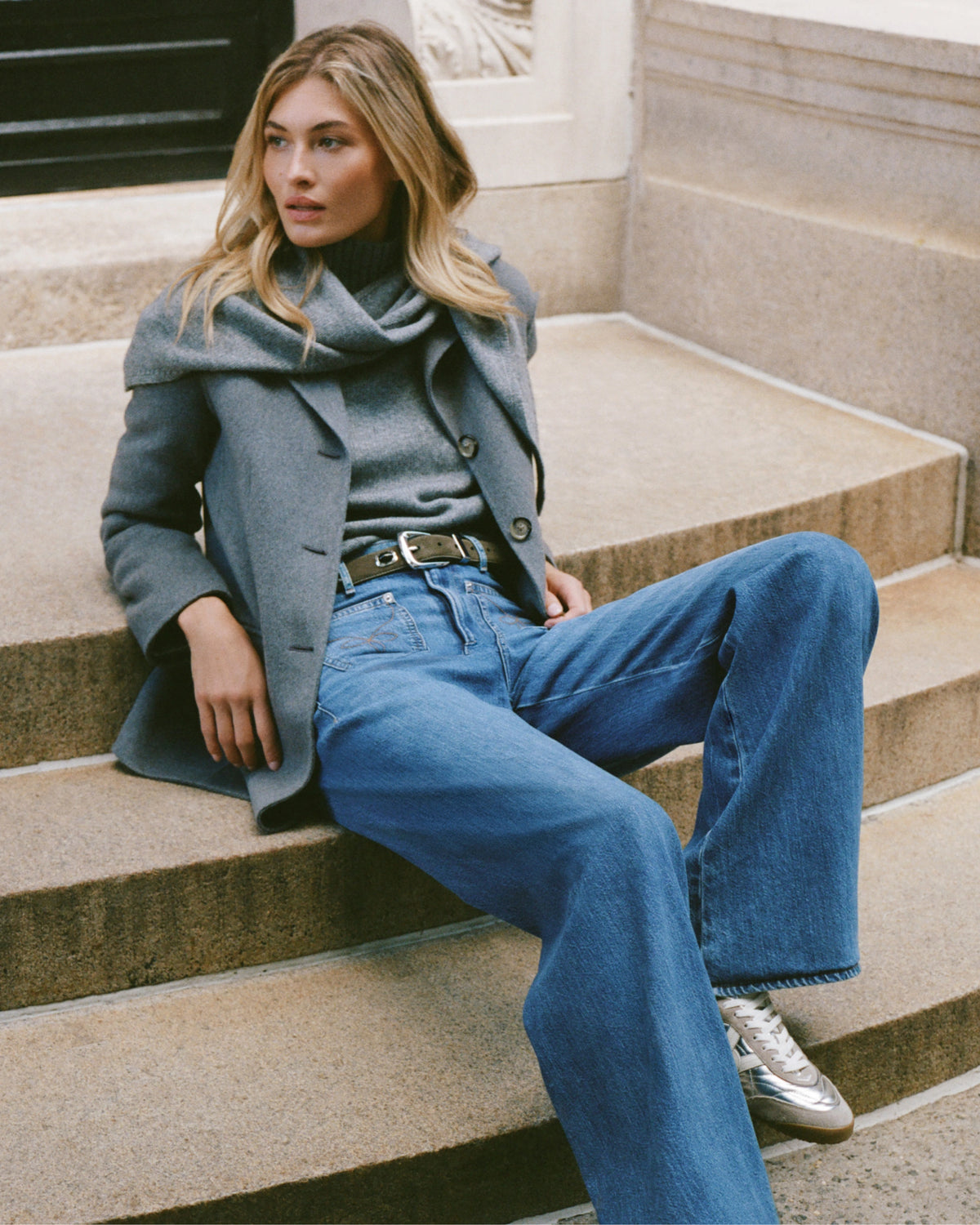 A woman in a gray coat and jeans sits on stone steps, with a blurred building in the background.