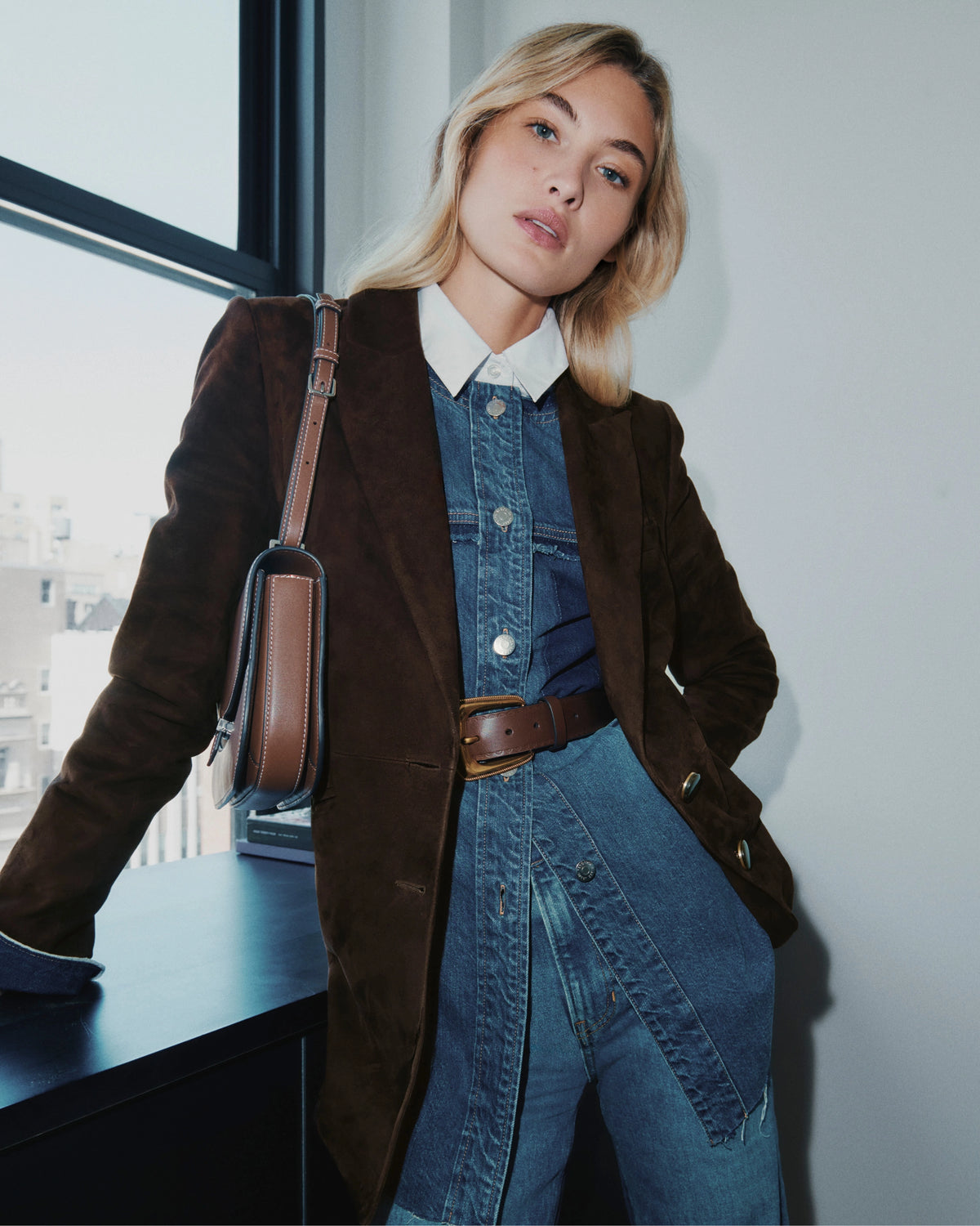 A blonde woman in a brown blazer and denim outfit stands in front of a window with a purse slung over her shoulder.