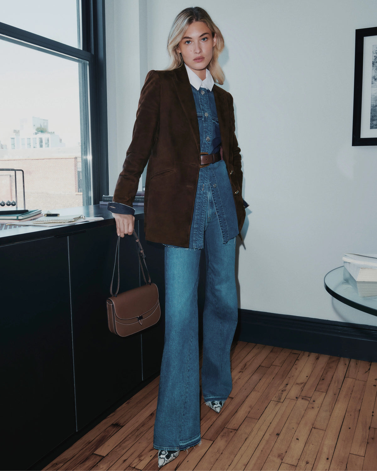 A woman in a brown blazer and denim outfit stands in a room with a window and a framed picture on the wall.