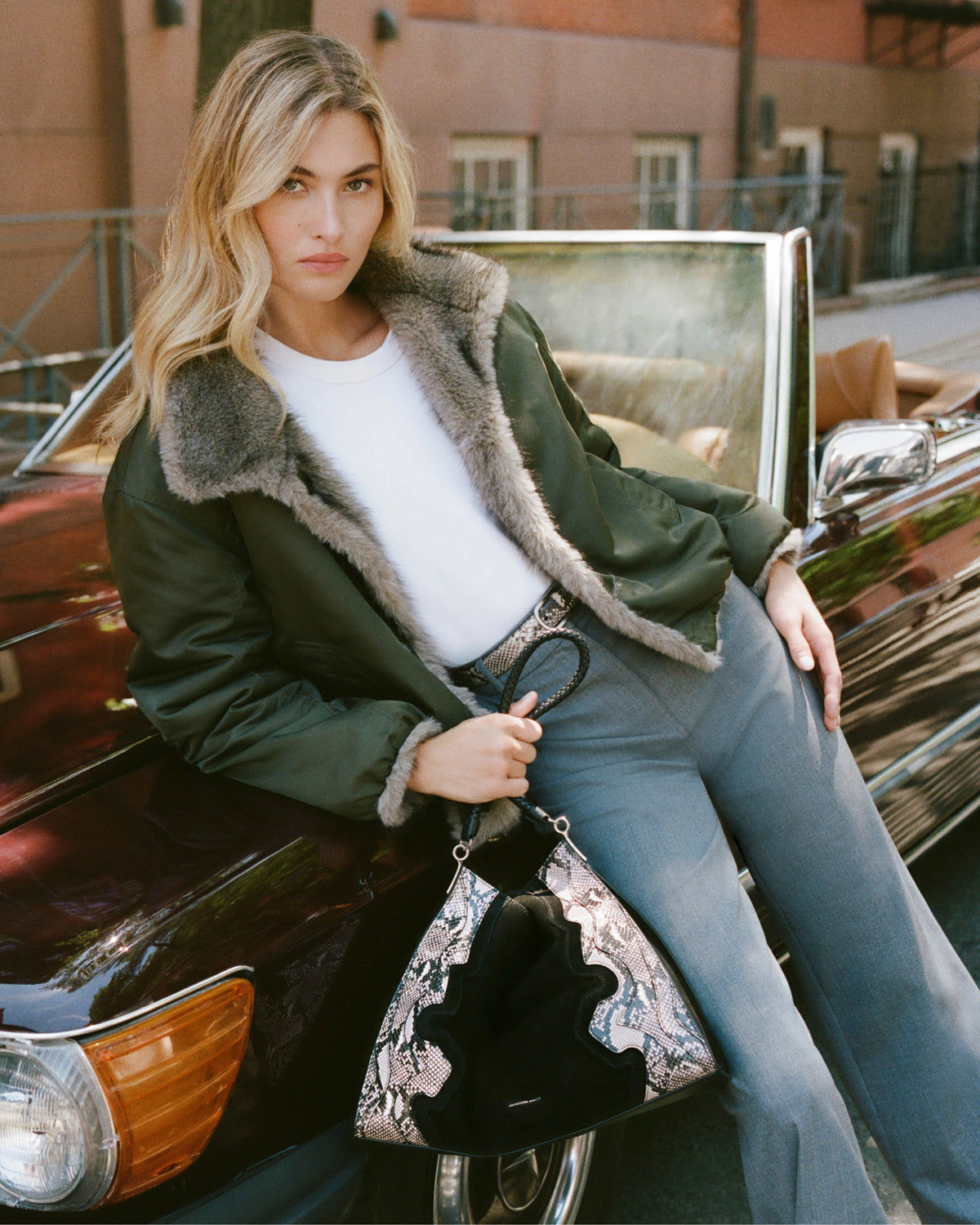A blonde woman in a fur-lined jacket and gray pants leans against a vintage convertible, holding a snakeskin-patterned handbag, with a brick building visible in the blurred background.