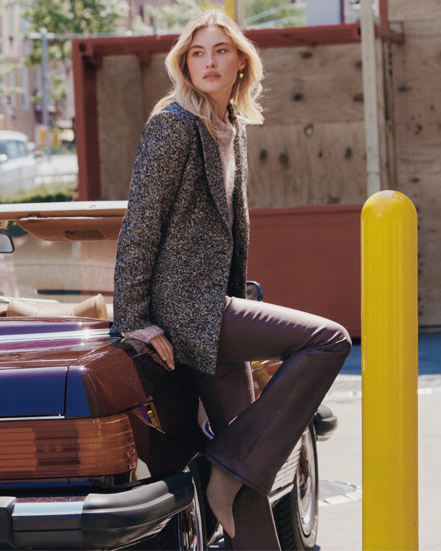 A blonde woman in a tweed blazer and flared pants leans against a vintage car, with a yellow bollard and urban buildings in the background.