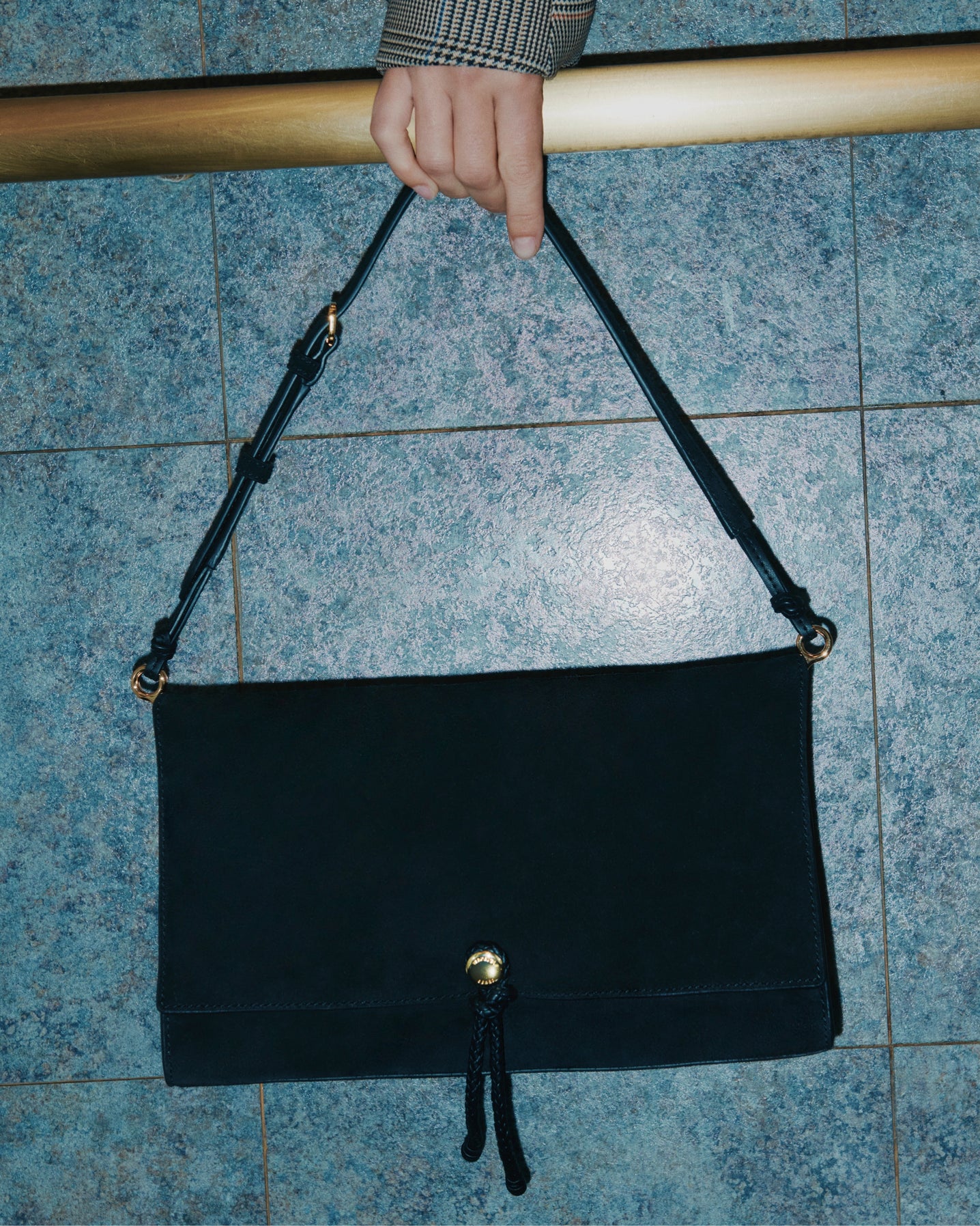 A hand holds a dark, rectangular handbag with a gold clasp in front of a blue tiled wall.