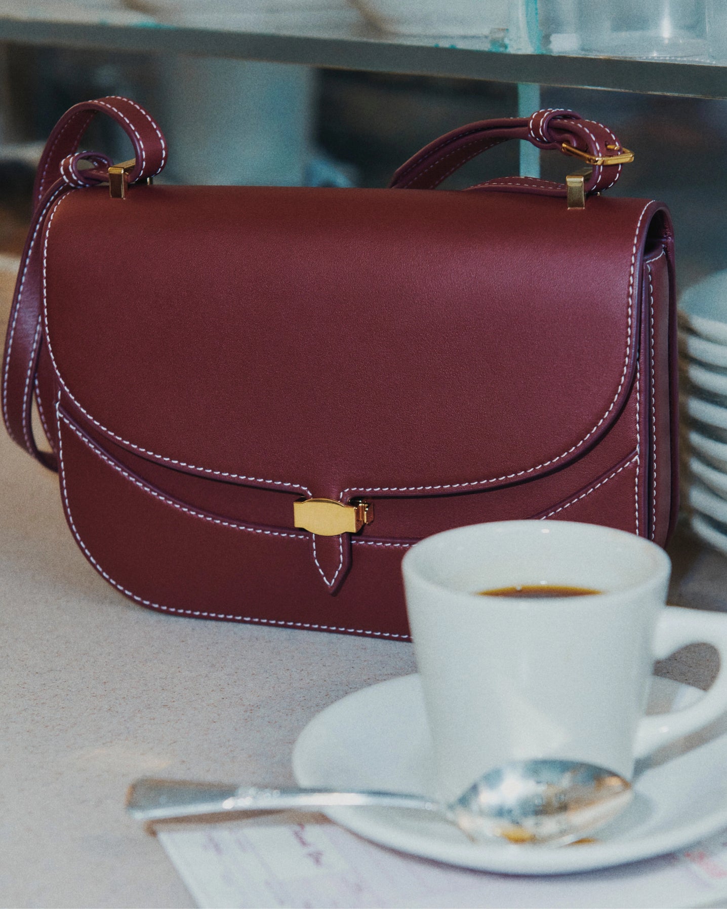 A burgundy leather handbag with white stitching sits on a counter next to a cup of coffee, with stacked plates visible in the blurred background.