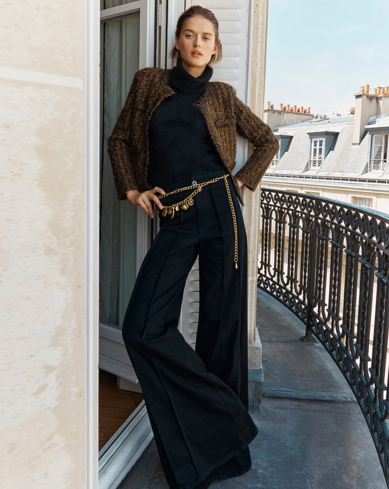 A woman in a black turtleneck, wide-leg trousers, and a textured jacket stands on a balcony overlooking Parisian rooftops.