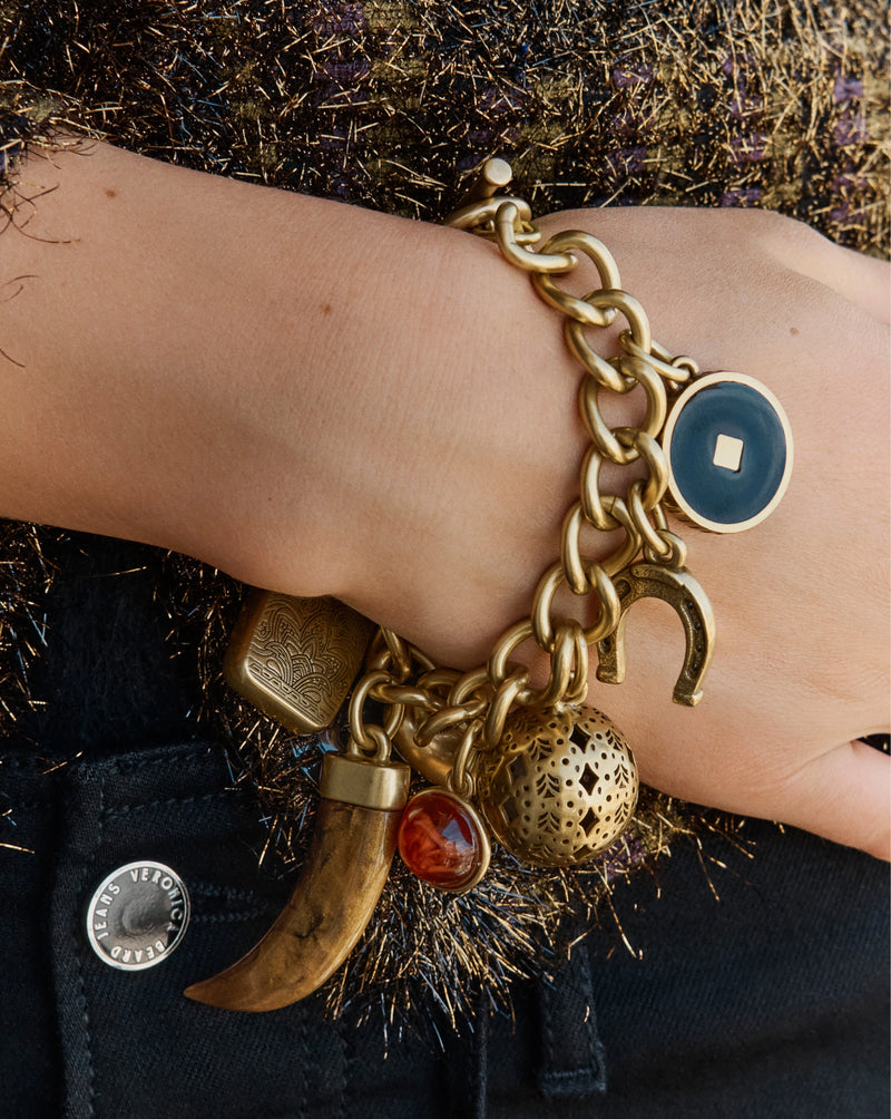 A gold charm bracelet with various ornaments rests on a person's wrist against a background of dark denim and shimmering gold fabric.