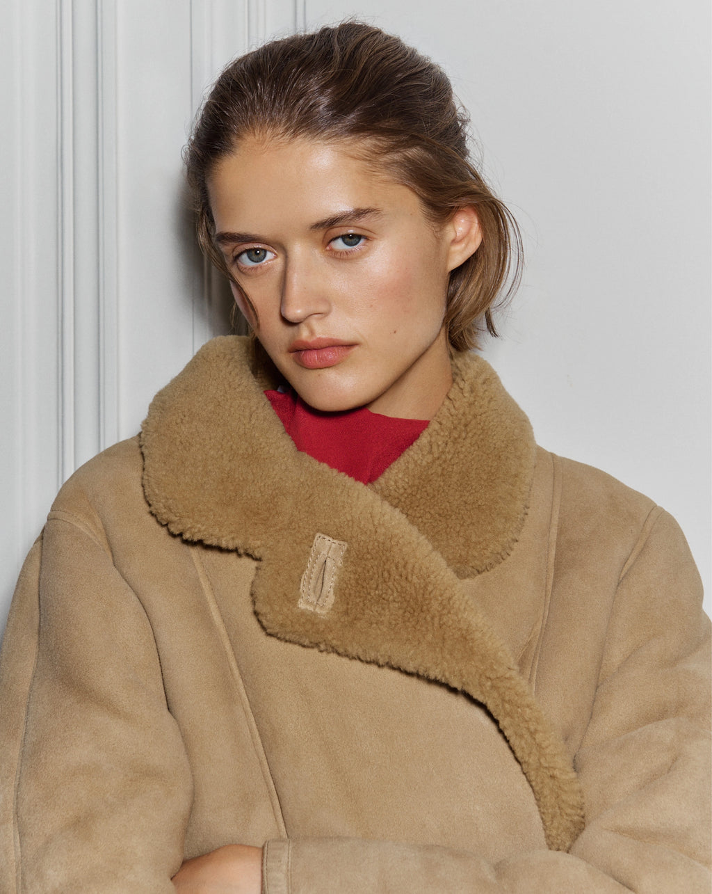 A woman wearing a tan shearling coat and a red shirt stands against a plain white wall.