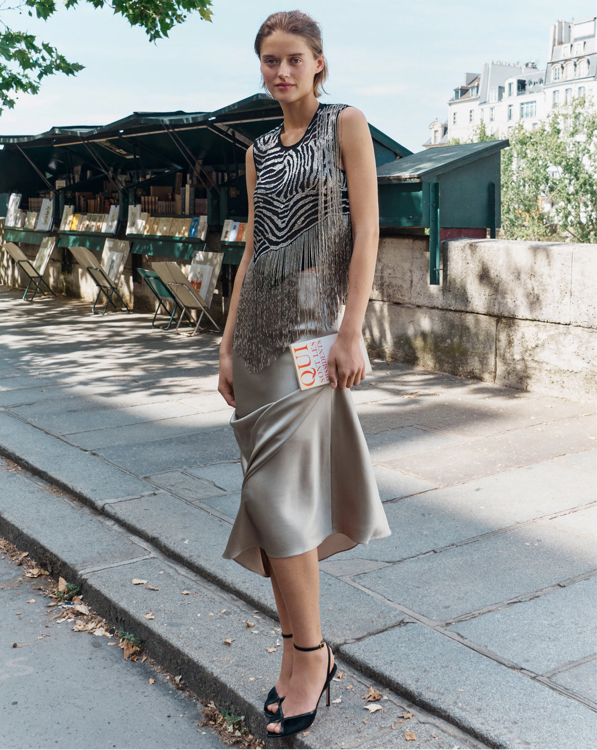 A woman in a fringed zebra-print top and satin skirt stands on a stone sidewalk in front of a row of outdoor book stalls.
