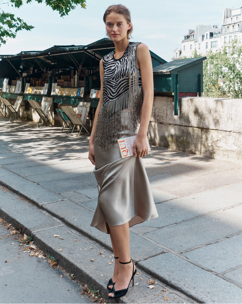 A woman in a fringed zebra-print top and satin skirt stands on a stone sidewalk in front of a row of outdoor book stalls.