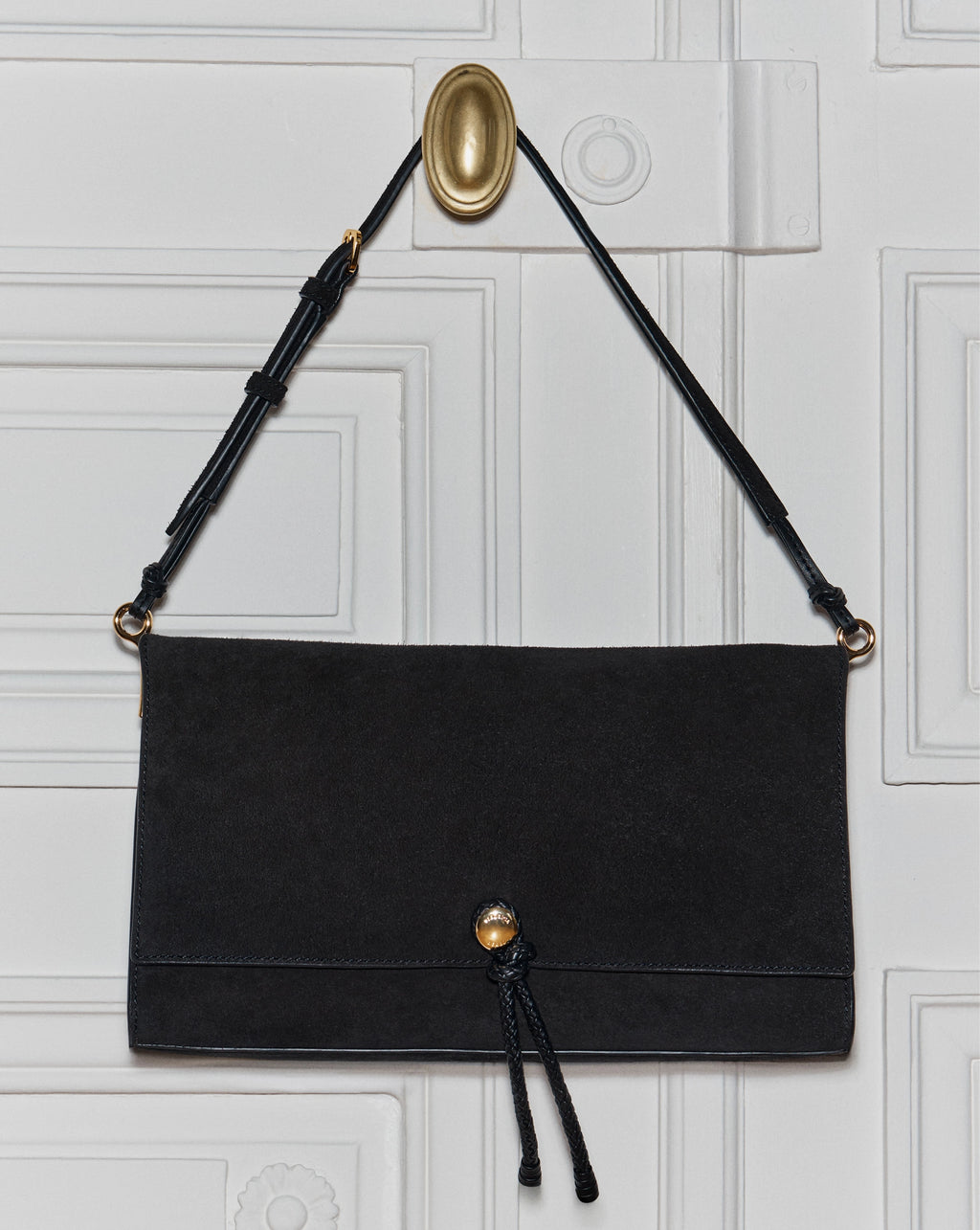 A black suede shoulder bag hangs from a gold hook on a white paneled door.