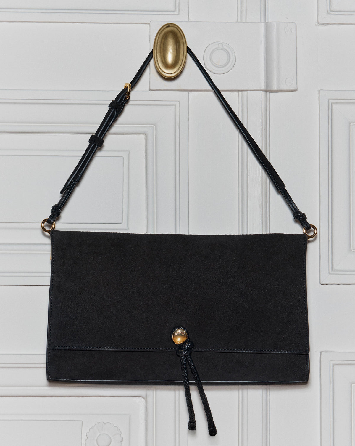 A black suede shoulder bag hangs from a gold hook on a white paneled door.