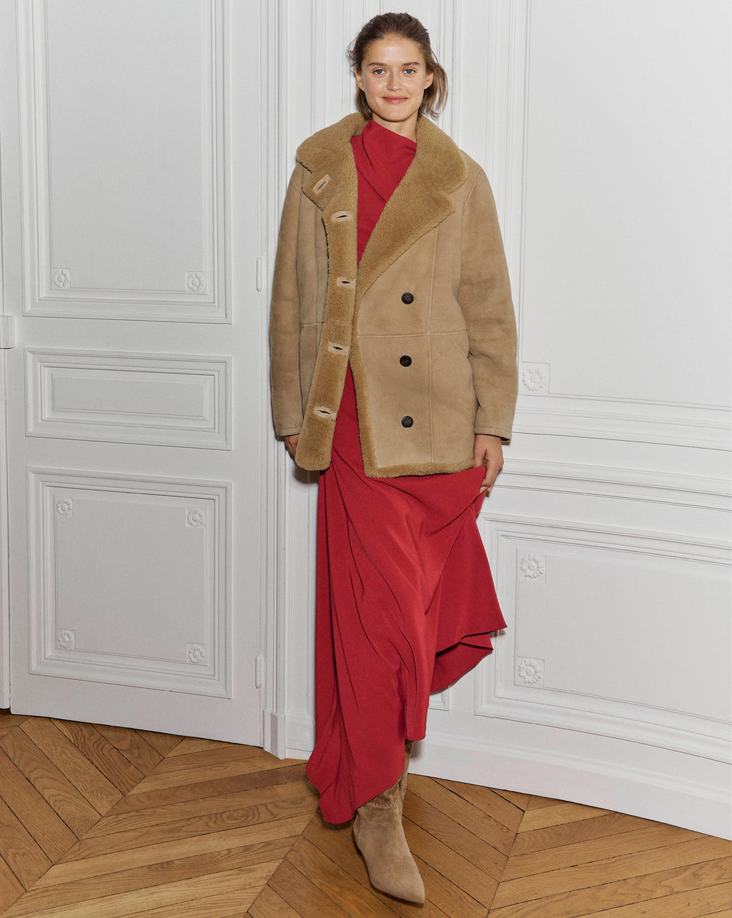 A woman wearing a tan shearling jacket over a long red dress stands against a white paneled wall on a herringbone wood floor.